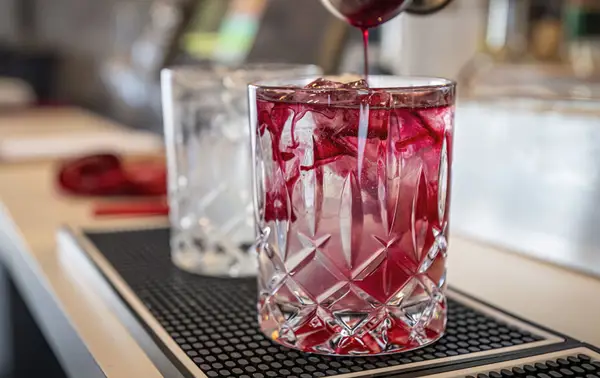 A cocktail is being prepared in a clear crystal glass filled with ice cubes. Red liquid is being poured, creating a swirling effect with the ice. In the background, another similar glass and blurred bar elements are visible.
