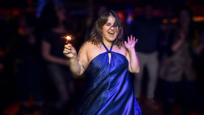 A smiling woman in a blue dress dances joyfully while holding a sparkler in a festive, well-lit setting, surrounded by people in the background.