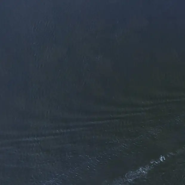 Vue aérienne sur le Louis Jolliet sur une vaste étendue d'eau bleu du St-Laurent. Le bateau avance en laissant derrière lui de légères ondulations. Il possède plusieurs ponts avec des structures visibles et des passagers à bord.