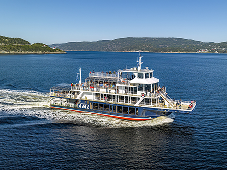 Le bateau "Le Grand Fleuve" de la compagnie AML navigue sur une eau calme, laissant une traînée derrière lui. Des passagers sont sur les ponts, admirant le paysage environnant composé de collines verdoyantes et d'un ciel bleu.