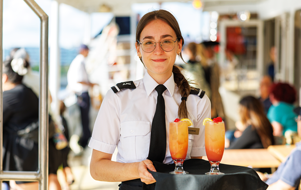 A smiling waitress in uniform holding a tray with two orange-colored cocktails on it, all aboard the AML Cavalier Maxim in Montreal.