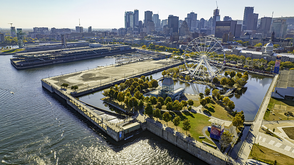 Aerial view of a large urban park surrounded by water, featuring a Ferris wheel, green spaces, and modern buildings in the background of a city.