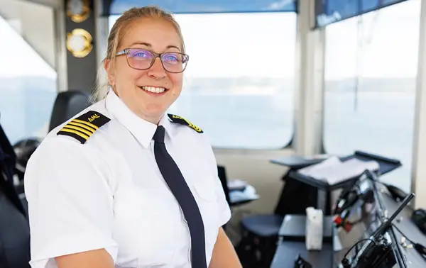 A person wearing a captain's uniform, including a white shirt with shoulder stripes and a black tie, is smiling with a lollipop in their mouth. They are in the control room of a vessel with navigation equipment around. A large window shows a body of water.