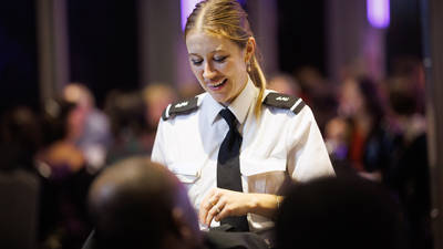 A waitress in a white uniform with a black tie smiles while serving a customer in a lively room with a warm atmosphere and dim lighting.
