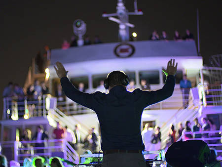 A DJ performs at a nighttime party on a boat, with colorful lights and a crowd dancing across multiple levels of the vessel.