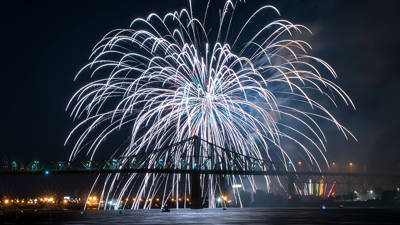 Stunning white fireworks burst in the night sky above an illuminated bridge, with shimmering reflections on the water and city lights in the background.