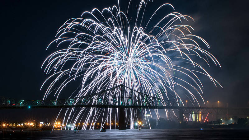 Stunning white fireworks burst in the night sky above an illuminated bridge, with shimmering reflections on the water and city lights in the background.