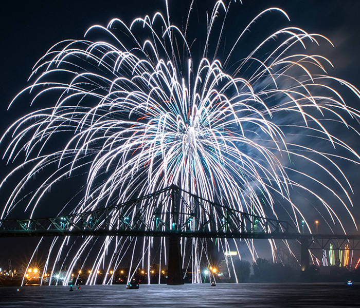 Stunning white fireworks burst in the night sky above an illuminated bridge, with shimmering reflections on the water and city lights in the background.