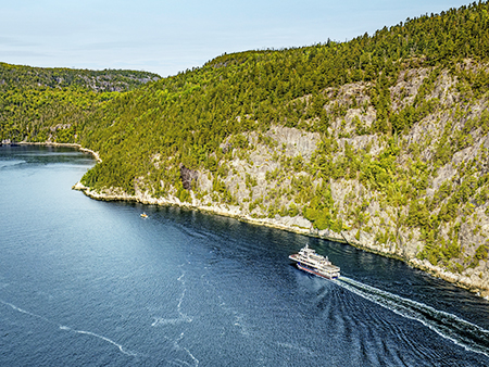 Vue a&#xE9;rienne d&#x27;un ferry naviguant &#xE0; travers une rivi&#xE8;re sinueuse bord&#xE9;e de for&#xEA;ts. Le navire laisse une tra&#xEE;n&#xE9;e dans l&#x27;eau bleu profond, tandis que des falaises rocheuses et des arbres denses et verdoyants longent les rives sous un ciel clair.