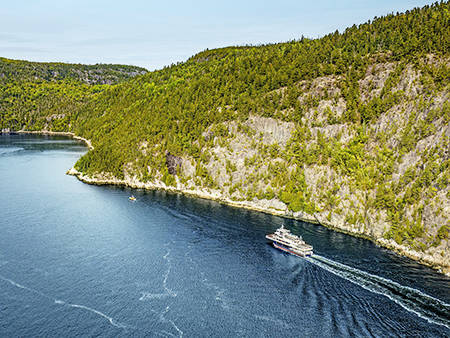 Aerial view of a ferry cruising through a winding, forested river. The vessel leaves a trail in the deep blue water, with rocky cliffs and dense green trees lining the riverbanks under a clear sky.