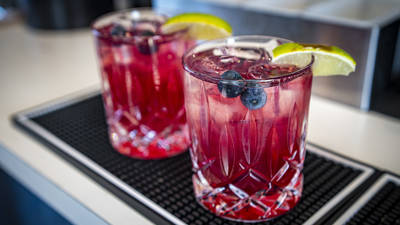 Two glasses of a vibrant red cocktail sit on a bar mat. Each drink is garnished with a lime wedge and a couple of blueberries. The glasses are intricately designed and contain ice cubes. The setting appears to be a bar or restaurant counter.