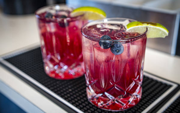 Two glasses of a vibrant red cocktail sit on a bar mat. Each drink is garnished with a lime wedge and a couple of blueberries. The glasses are intricately designed and contain ice cubes. The setting appears to be a bar or restaurant counter.