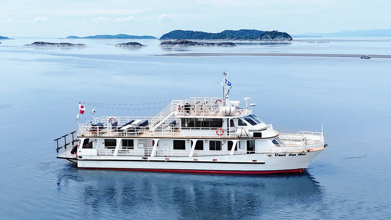 Le bateau "Vent des Îles" navigue tranquillement sur des eaux calmes entourées d'îles et d'une légère brume à l'horizon. Le ciel est dégagé et lumineux, créant une ambiance paisible.