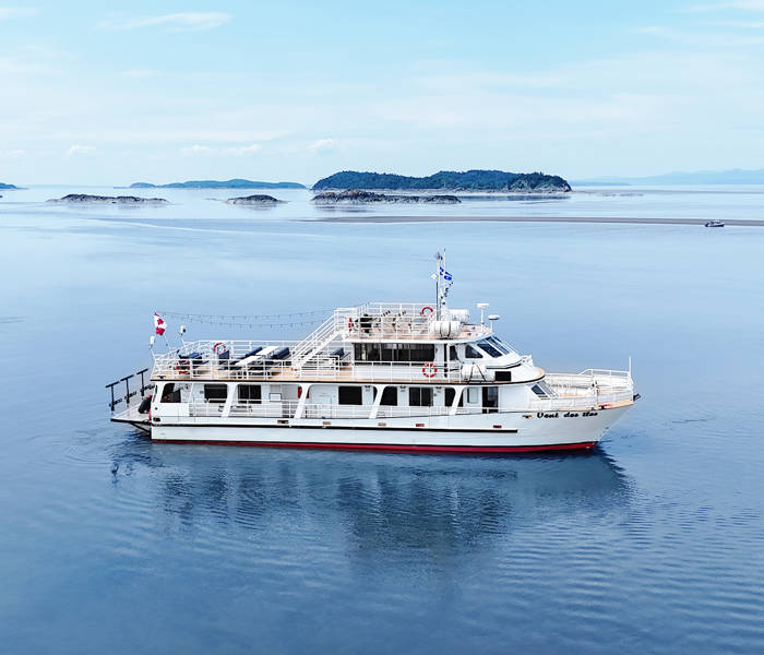 Le bateau "Vent des Îles" navigue tranquillement sur des eaux calmes entourées d'îles et d'une légère brume à l'horizon. Le ciel est dégagé et lumineux, créant une ambiance paisible.