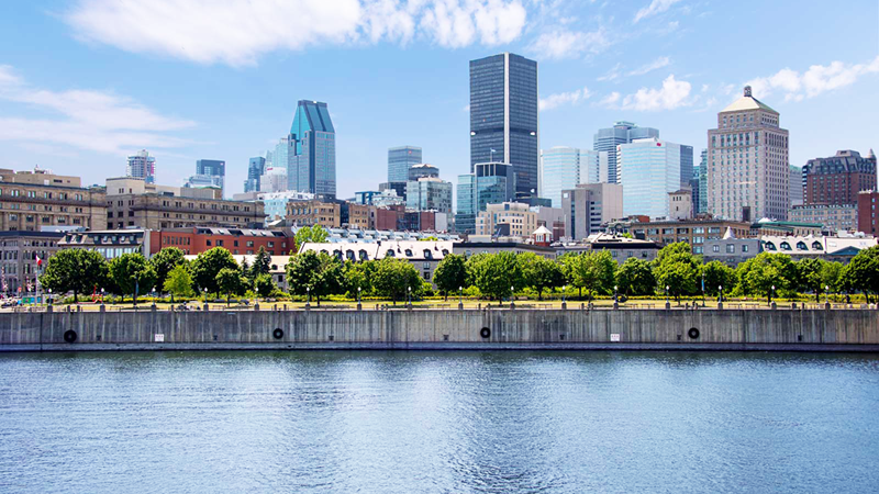 The Montreal skyline with modern buildings under a blue sky, with the St. Lawrence River in the foreground.