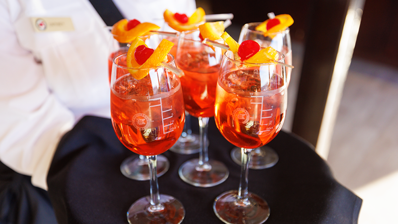 A close-up of a server's tray with 6 glasses of orange-colored Spritz, each garnished with an orange wedge and a cherry.
