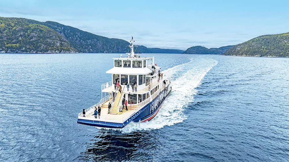 Un bateau de croisière navigue sur une étendue d'eau calme, laissant une traînée blanche derrière lui. Des passagers se tiennent sur le pont, admirant le paysage environnant composé de collines verdoyantes sous un ciel légèrement couvert.