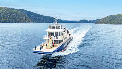 A cruise ship sails on calm waters, leaving a white wake behind it. Passengers stand on the deck, enjoying the surrounding scenery of green hills under a slightly overcast sky.