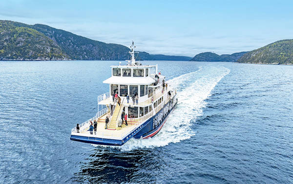 A cruise ship sails on calm waters, leaving a white wake behind it. Passengers stand on the deck, enjoying the surrounding scenery of green hills under a slightly overcast sky.