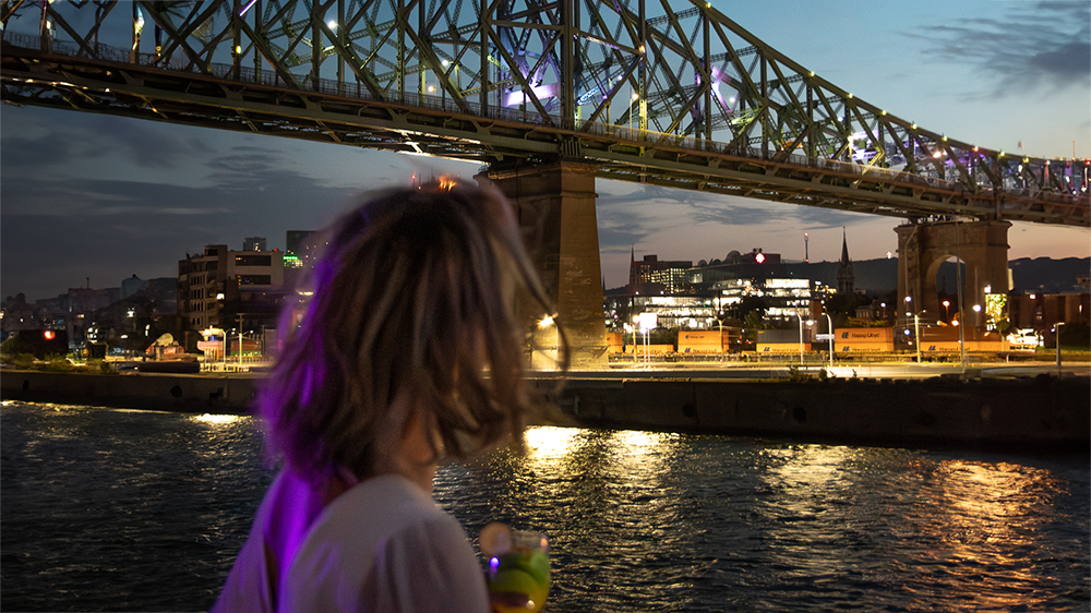 A woman viewed from behind holding a drink, admiring a lit-up bridge spanning a river, with city lights and a twilight sky in the background.