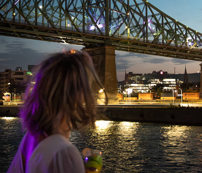A woman viewed from behind holding a drink, admiring a lit-up bridge spanning a river, with city lights and a twilight sky in the background.