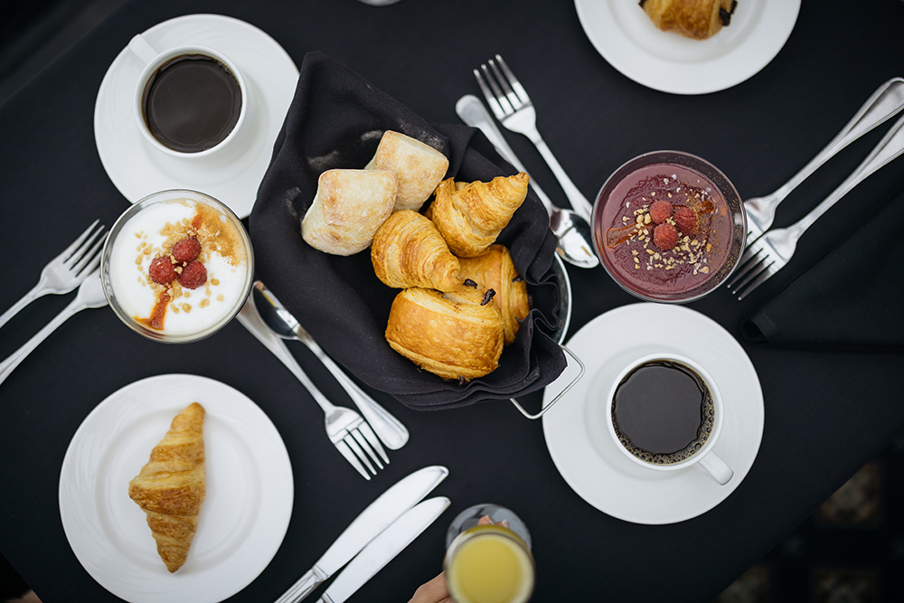 Vue aérienne d'une table de petit-déjeuner comprenant des croissants, des petits pains dans un panier, des tasses de café, un verre de smoothie garni de granola et de baies, ainsi qu'un parfait au yaourt et aux framboises. Les couverts sont soigneusement disposés autour des plats.