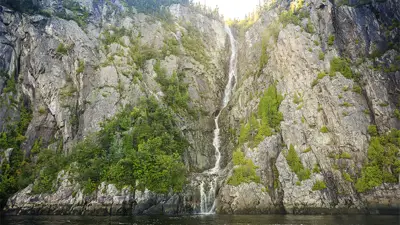A tall waterfall cascades down a steep, rocky cliff surrounded by lush green vegetation. The water flows into a calm body of water at the base, with sunlight illuminating the top of the cliff.