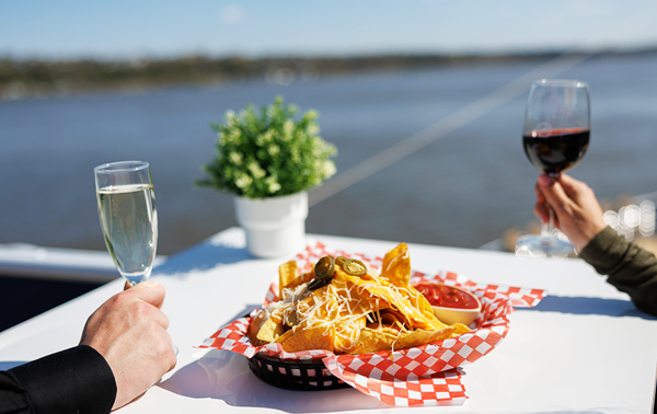 Two people sitting at a table with a view of the river, one holding a glass of wine and the other a flute of champagne, sharing a basket of nachos.