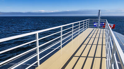 View from the deck of a ship, featuring white railings casting shadows on the beige deck. The vast ocean stretches out under a clear blue sky, with a distant horizon visible. Red and blue life preservers are attached to the railing.