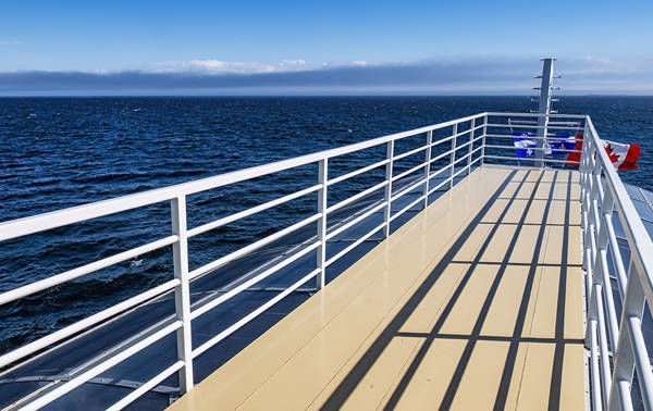 View from the deck of a ship, featuring white railings casting shadows on the beige deck. The vast ocean stretches out under a clear blue sky, with a distant horizon visible. Red and blue life preservers are attached to the railing.