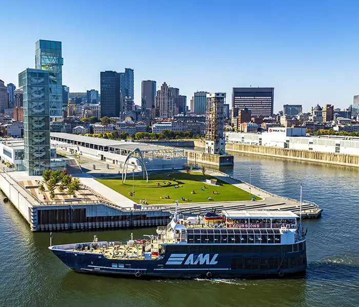 Vue aérienne du navire AML Cavalier Maxim stationné devant la Tour du Port de Montréal de jour avec vue sur la skyline ensoleillée de la ville en arrière plan.