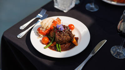 A gourmet dish served on a white plate, featuring a filet of meat with sauce, vegetables, a lobster tail, and a floral garnish, placed on an elegant table with a black tablecloth.
