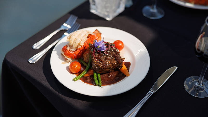 A gourmet dish served on a white plate, featuring a filet of meat with sauce, vegetables, a lobster tail, and a floral garnish, placed on an elegant table with a black tablecloth.