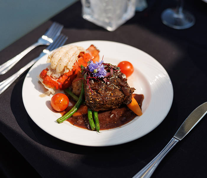A gourmet dish served on a white plate, featuring a filet of meat with sauce, vegetables, a lobster tail, and a floral garnish, placed on an elegant table with a black tablecloth.