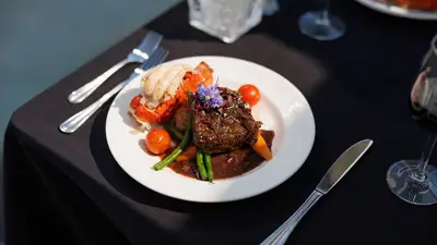 Un plat gastronomique servi sur une assiette blanche, comprenant un filet de viande nappé de sauce, des légumes, une queue de homard et une garniture florale, présenté sur une table élégante avec une nappe noire.