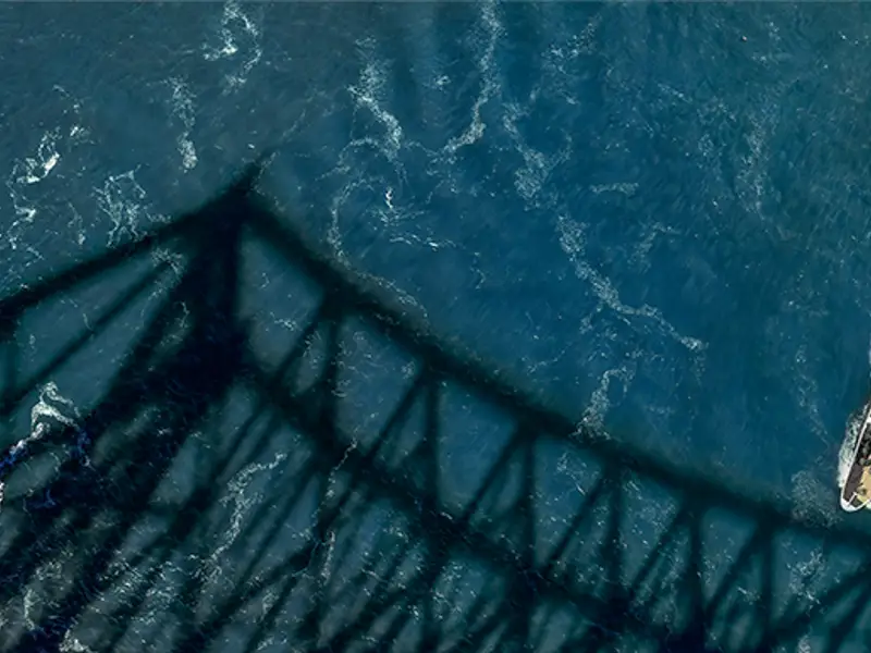 Aerial view of a boat sailing through the blue waters of the Saint Lawrence under a large bridge casting a shadow. The shadow creates a striking pattern on the surface of the water.