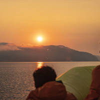 Passengers wearing orange jackets watch a sunset over calm waters. In the distance, mist-covered hills add a sense of mystery to the scene. The sky is painted with golden and orange hues.