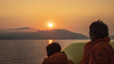 Passengers wearing orange jackets watch a sunset over calm waters. In the distance, mist-covered hills add a sense of mystery to the scene. The sky is painted with golden and orange hues.