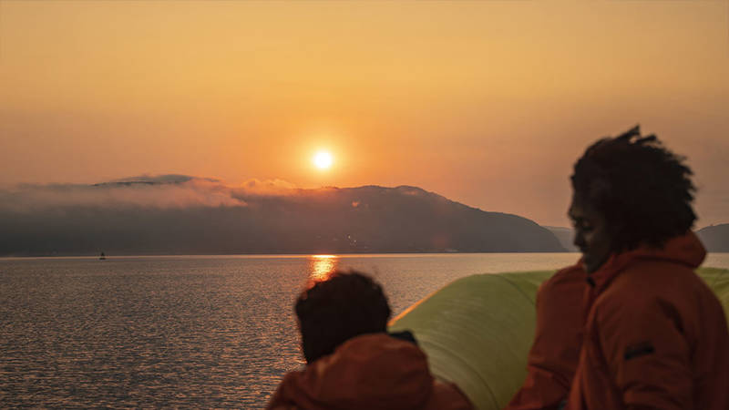 Passengers wearing orange jackets watch a sunset over calm waters. In the distance, mist-covered hills add a sense of mystery to the scene. The sky is painted with golden and orange hues.