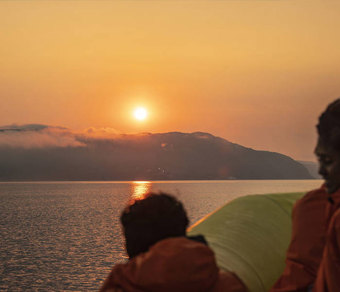 Passengers wearing orange jackets watch a sunset over calm waters. In the distance, mist-covered hills add a sense of mystery to the scene. The sky is painted with golden and orange hues.