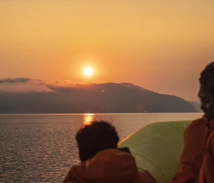 Des passagers vêtus de manteaux orange regardent un coucher de soleil sur une étendue d'eau calme. À l'horizon, des collines enveloppées de brume ajoutent une touche de mystère à la scène. Le ciel est teinté de nuances dorées et orangées.