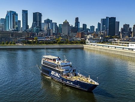 The ship "Cavalier Maxim" from AML cruises along a river bordered by a modern cityscape with tall skyscrapers in the background. The sky is clear, and daylight brightens the urban landscape and water.