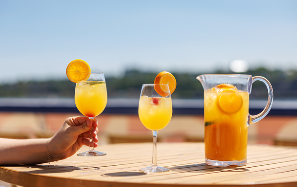 A close-up of a hand holding a glass of orange sangria, with another glass and a pitcher of sangria served on a wooden table on one of the terraces of the AML Cavalier Maxim in Montreal.