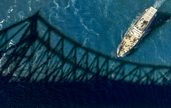 Aerial view of the Cavalier Maxime sailing on the sparkling St. Lawrence River, with the shadow of the Jacques Cartier Bridge in the foreground.
