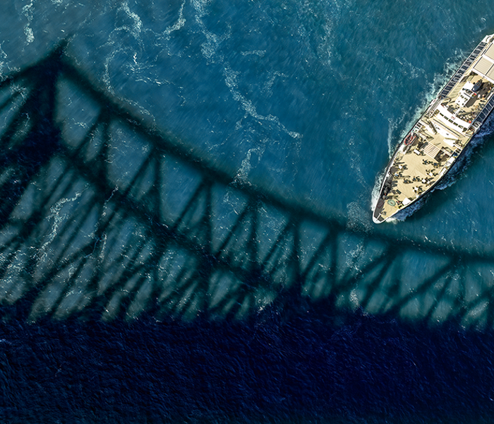 Aerial view of the Cavalier Maxime sailing on the sparkling St. Lawrence River, with the shadow of the Jacques Cartier Bridge in the foreground.