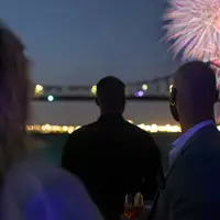 Un feu d'artifice coloré éclate dans le ciel nocturne près du pont Jacques-Cartier et de la tour de l'Horloge, avec des reflets sur le fleuve et une foule observant depuis le quai.