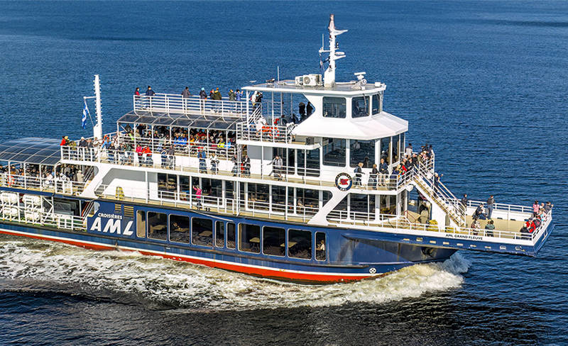 The ship "Le Grand Fleuve" from AML cruises sails on calm waters, leaving a wake behind. Passengers are on the decks, enjoying the surrounding scenery of green hills and a clear blue sky.