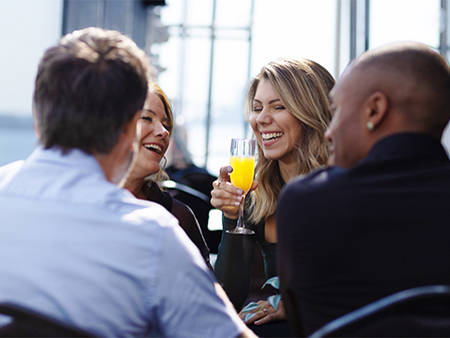 A group of four friends sharing a joyful moment in a restaurant near the water, with a smiling woman holding a glass of mimosa.