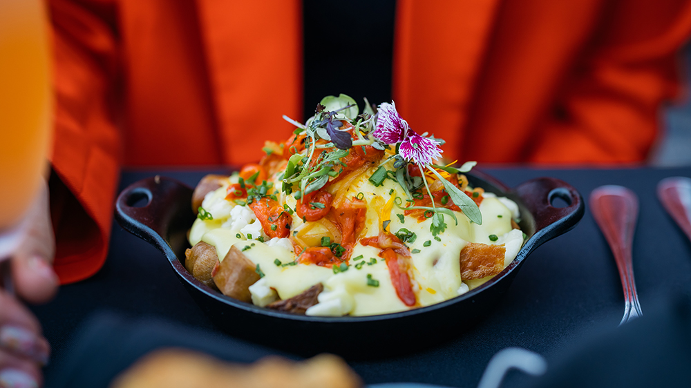 A delicious dish of roasted potatoes topped with hollandaise sauce, garnished with vegetables, fresh herbs, and edible flowers, served in a cast-iron skillet.