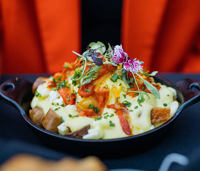 A delicious dish of roasted potatoes topped with hollandaise sauce, garnished with vegetables, fresh herbs, and edible flowers, served in a cast-iron skillet.